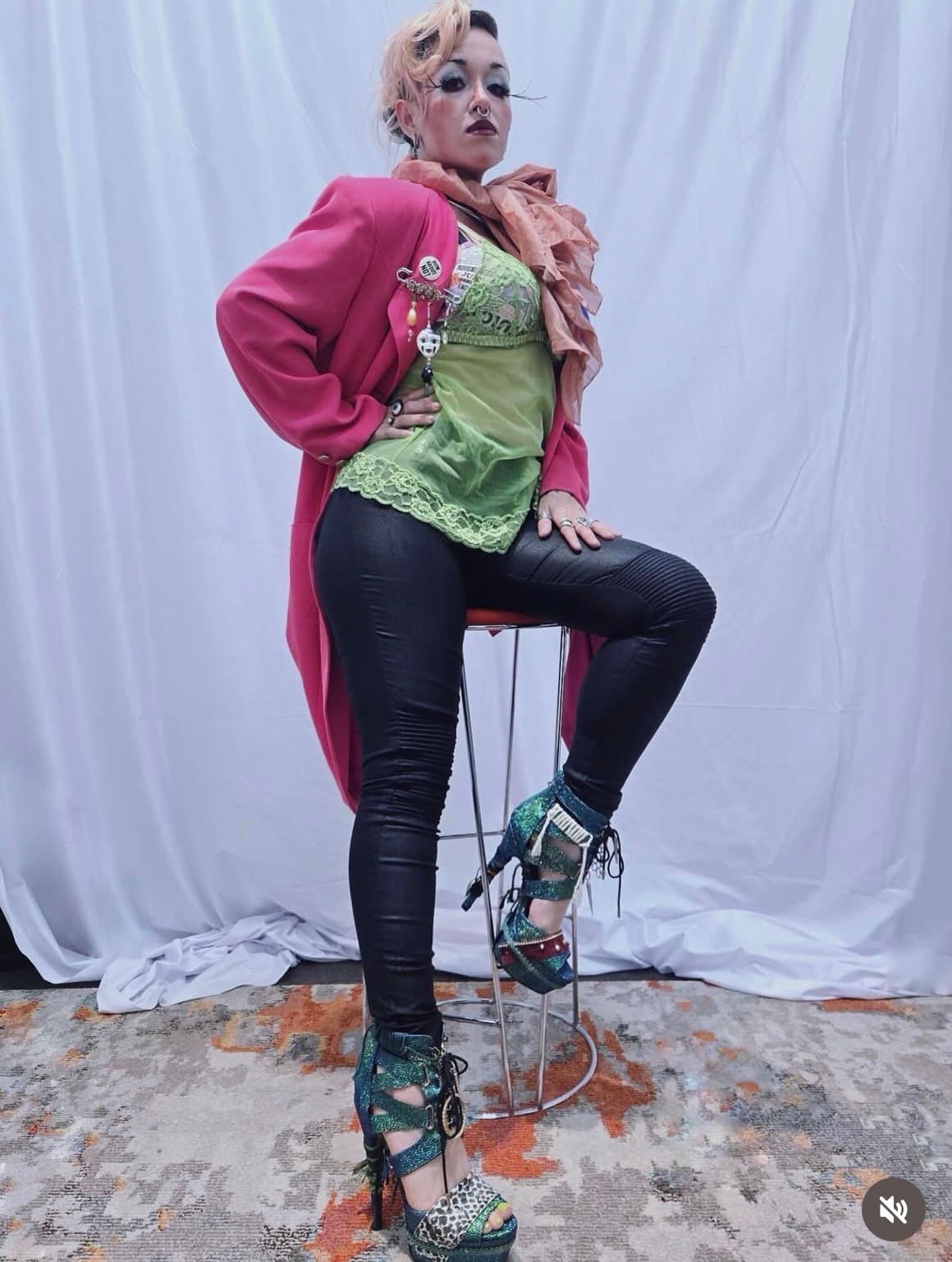 Bunny posing on a stool in a coral blazer, lime lace bustier, leather pants, and towering iridescent platform heels.