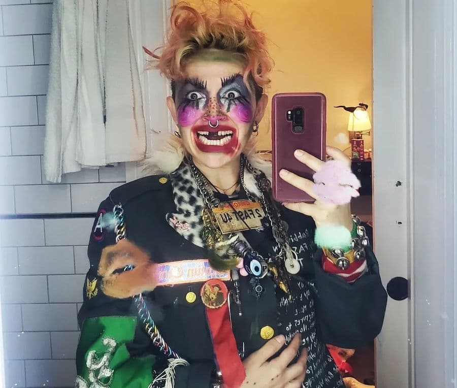 Bunny in a mirror selfie wearing a patched leather jacket, fox stole, and bold clown-style makeup in magenta and silver.