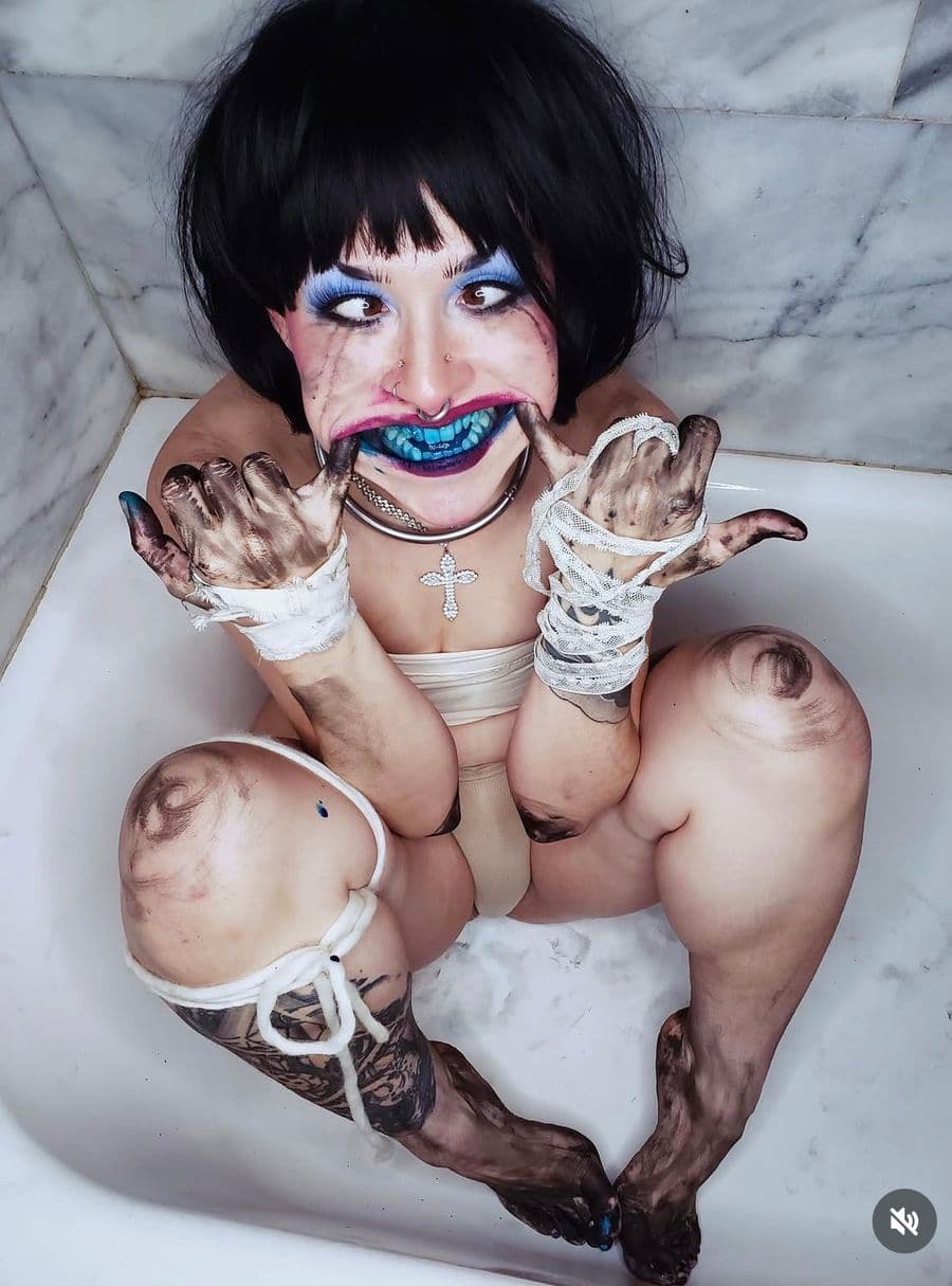 Close shot of Bunny in a porcelain tub, electric blue lips and lids, hands wrapped in linen and stained with black pigment.